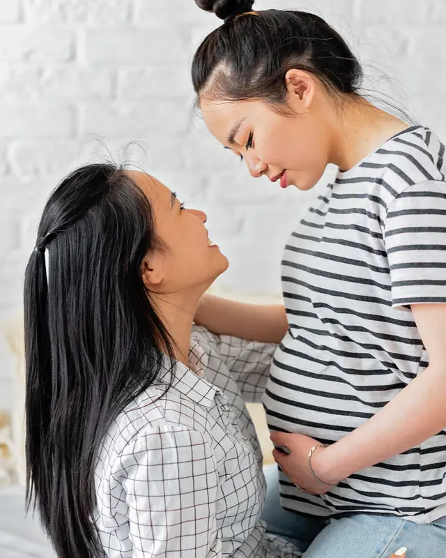 Two women share a tender look, one gently holding her pregnant belly