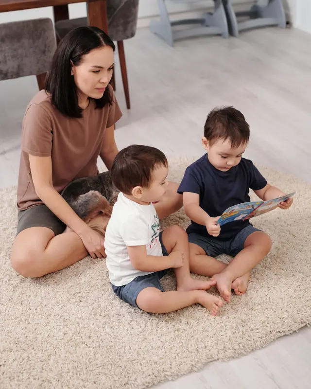A mother sits on a rug with her twin toddlers looking at a picture book