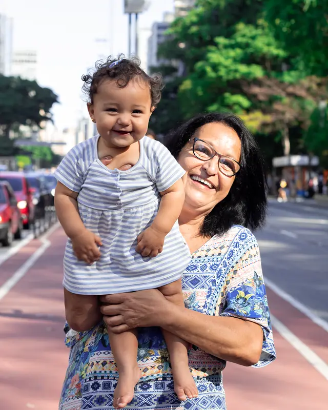 A smiling woman with glasses lifts a laughing baby on a sunny city street