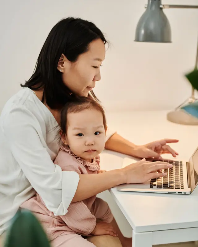 A mother types on a laptop at her desk with her toddler sitting on her lap